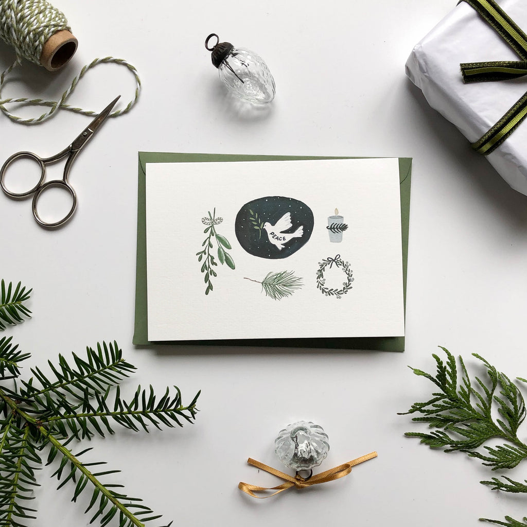 hand illustrated peace dove christmas card on table with glass baubles, pine branches & gift wrap