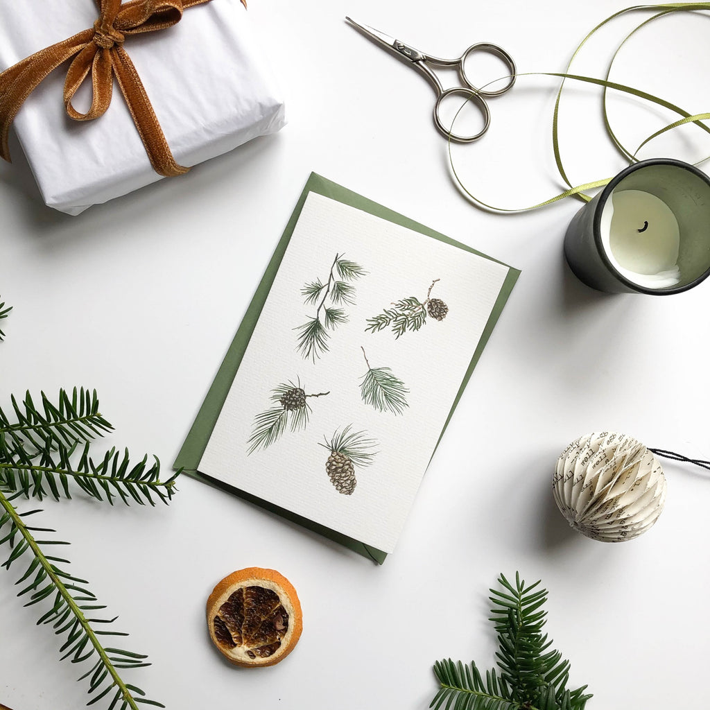 christmas card with hand illustrated christmas pine foliage on table with scissors & gift wrapped box & candle